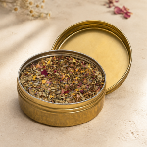 Open brass tin filled with Tobacco Blend and colorful dried botanicals, displayed with its lid on a warm natural surface.