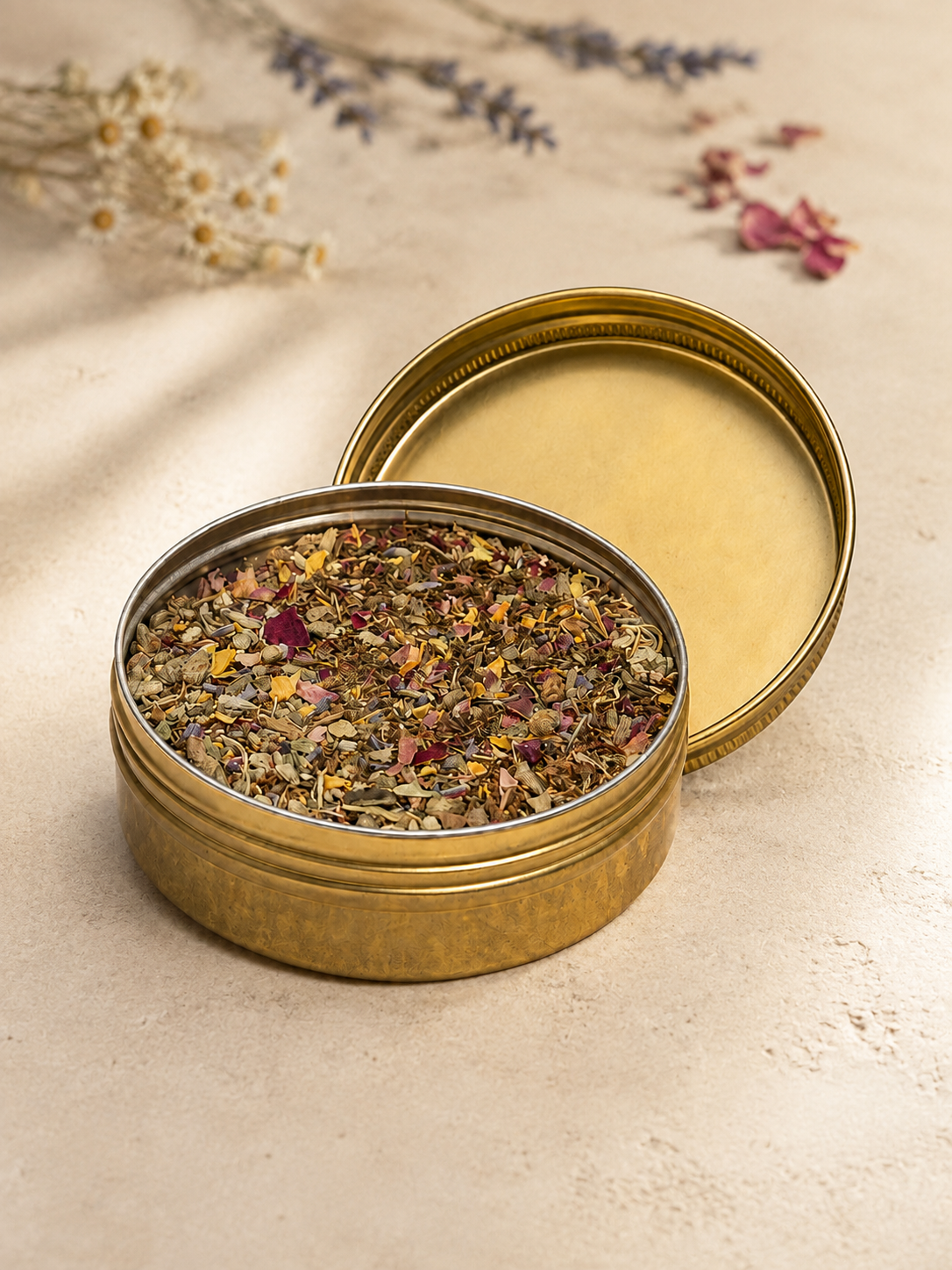 Open brass tin filled with Tobacco Blend and colorful dried botanicals, displayed with its lid on a warm natural surface.